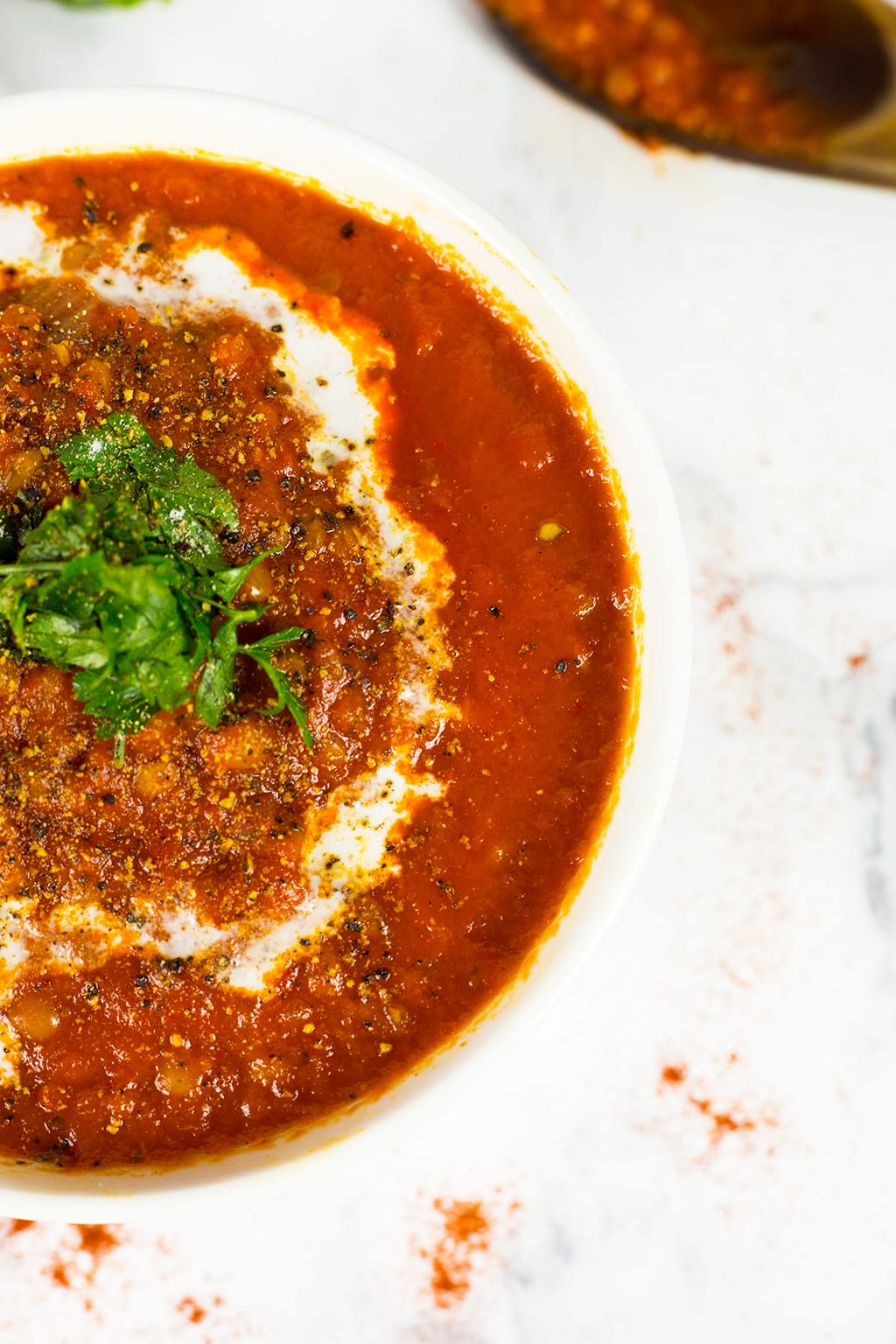 Roasted Red Pepper, Tomato & Lentil Soup with a swirl of coconut cream and parsley on top. Bowl on a white background with a wooden sppon behind it.