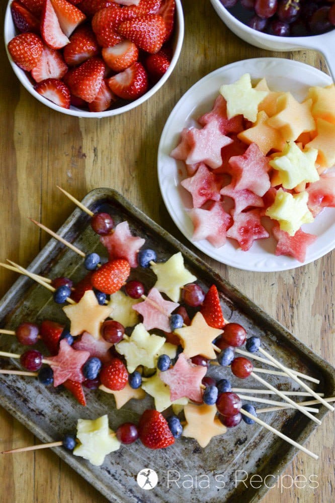 Fruit kabobs on baking tray with bowl of fruit to the side, some are cut into star shapes.