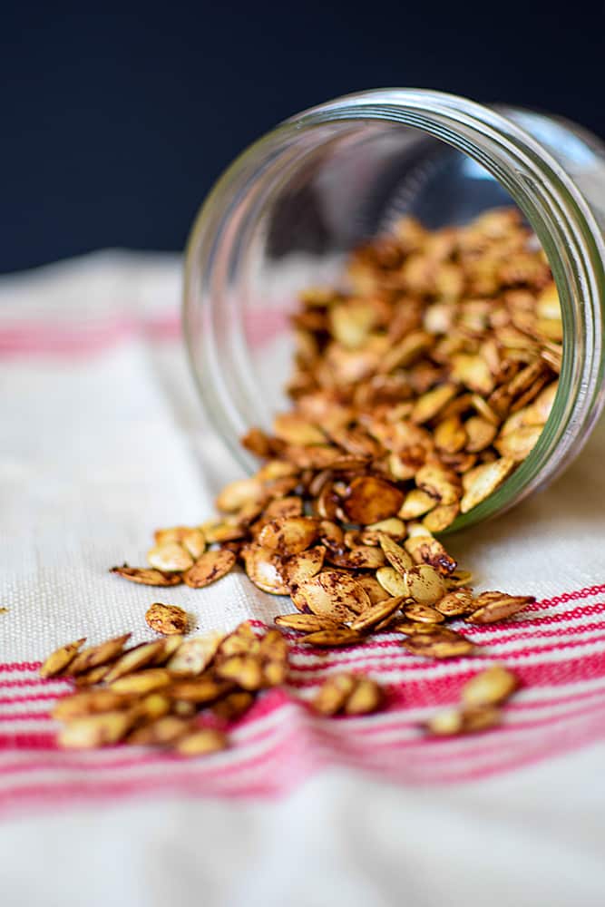 Roasted pumpkin seeds spilling out of a jar on white and red cloth.