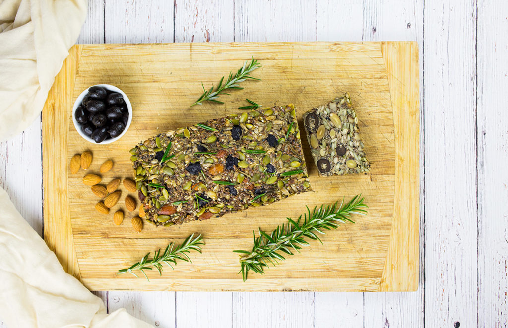 Paleo Nut and Seed Bread on chopping board with rosemary sprigs, bowl of olives and almonds beside it on white background.