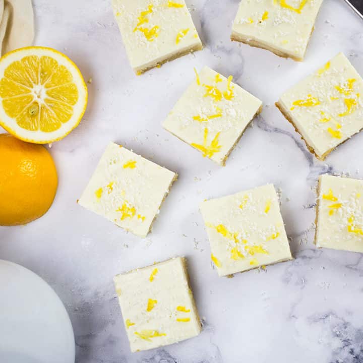 Birds eye view of creamy lemon squares on marble background with teapot and sliced lemon to the side.