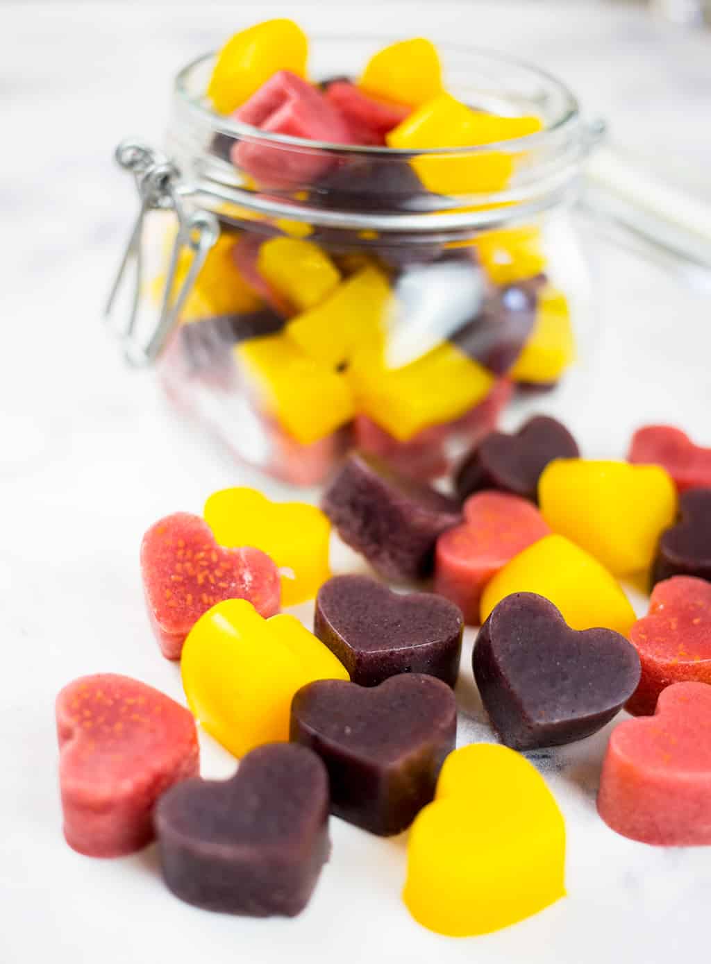 Mango gummies, raspberry gummies and blueberry gummies spilling out of glas jar on white background.