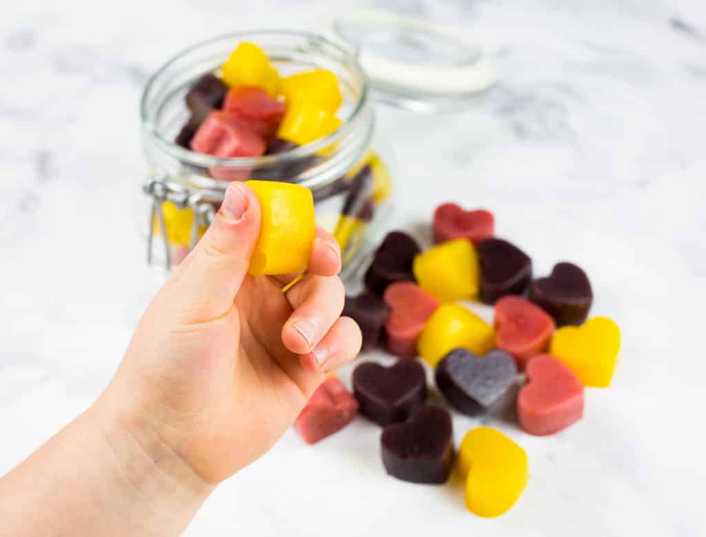 Picture of toddlers hand holding up a mango gummie in front of jar.