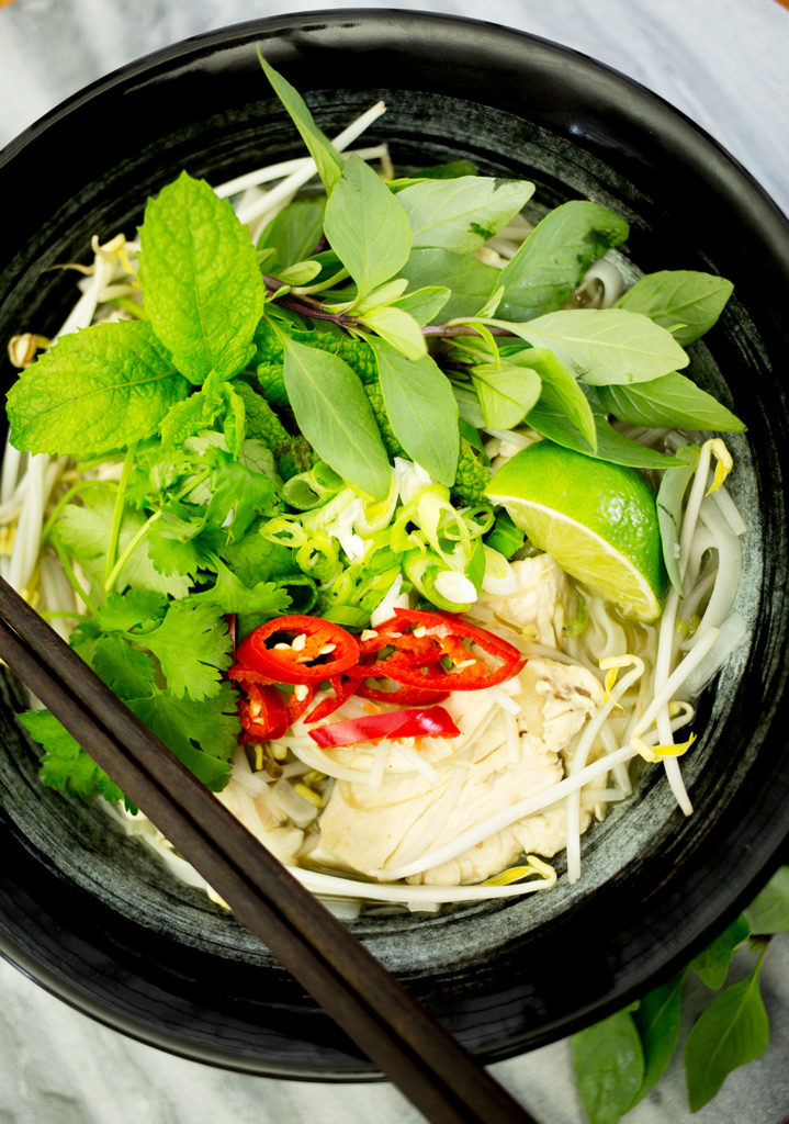 Birds eye view of Chicken Pho with fresh herbs, chili on top in black bowl on marble background.