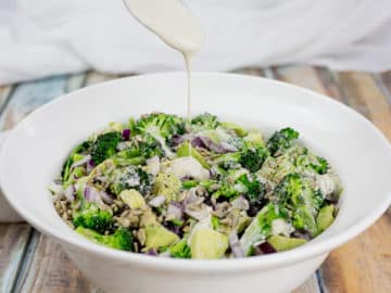 Healthy broccoli salad with cashew dressing being poured over it with a spoon, on wooden background with white cloth at the back.