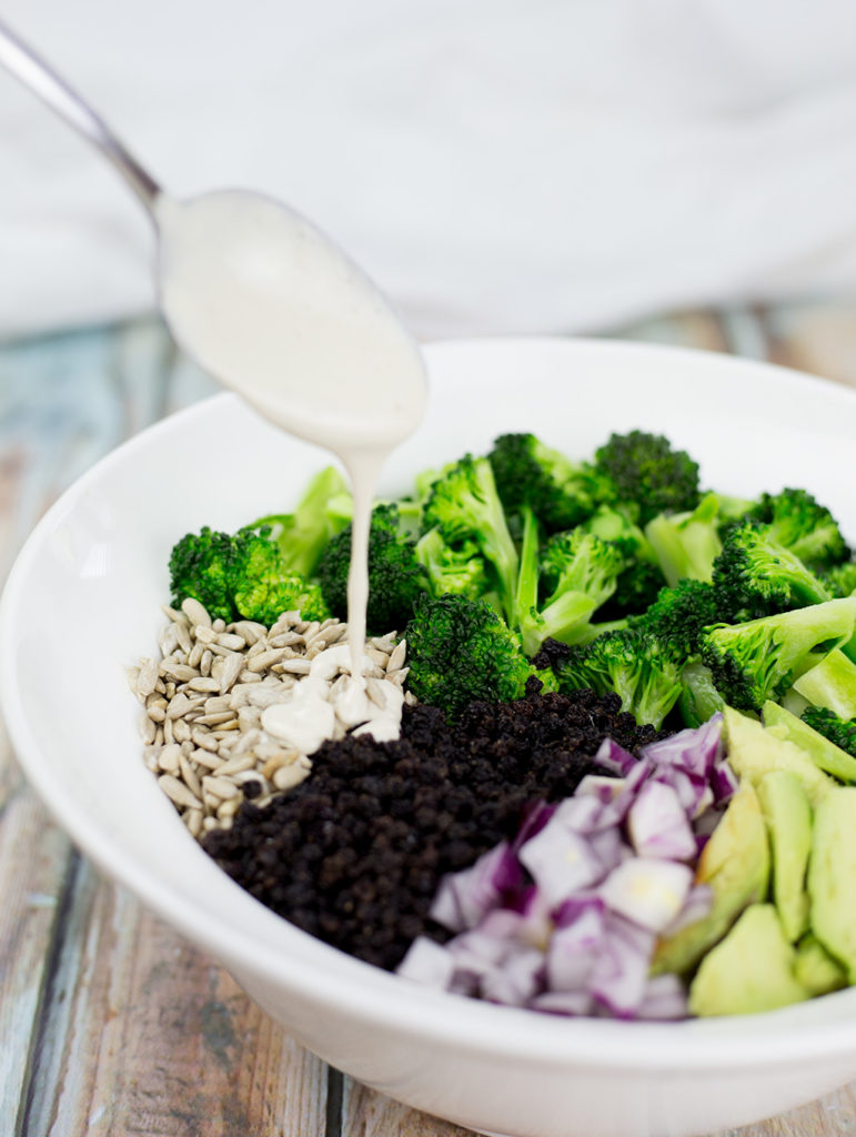 Healthy Broccoli Salad with veggies and creamy dressing being poured over the top with a spoon. The broocli, sunflower seeds, onion, and avocado is in seprate sections of the bowl.