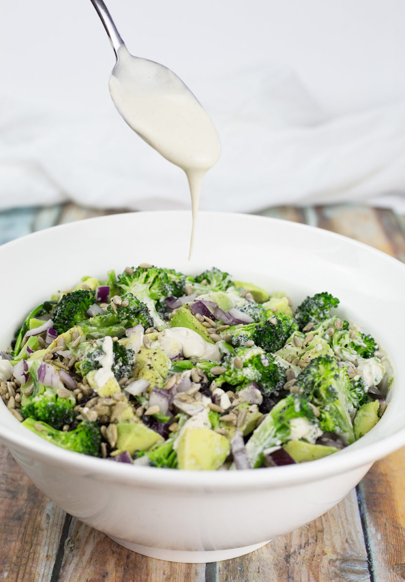 Dairy free dressing being poured over broccoli salad in a white bowl on a wooden background.