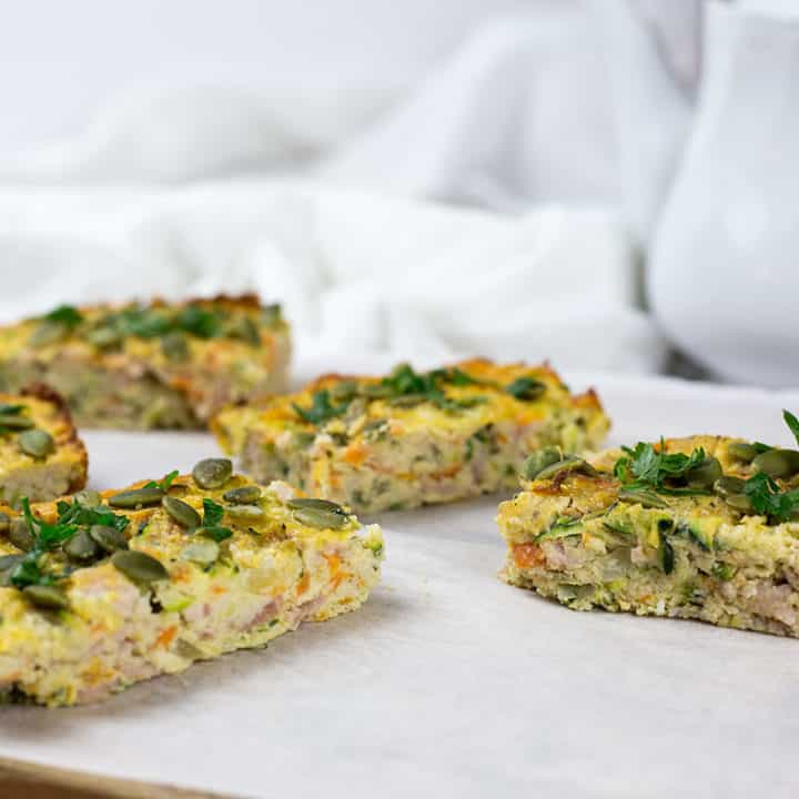 Close up of zucchini and bacon slice on chopping board with white parchment paper and teapot in the background.