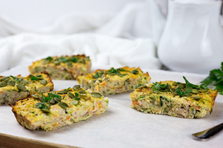 Close up of zucchini and bacon slice on chopping board with white parchment paper and teapot in the background.