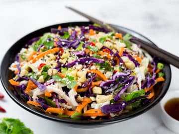 Asian slaw in black bowl with asian style dressing, chillis and coriander beside it on a white background.