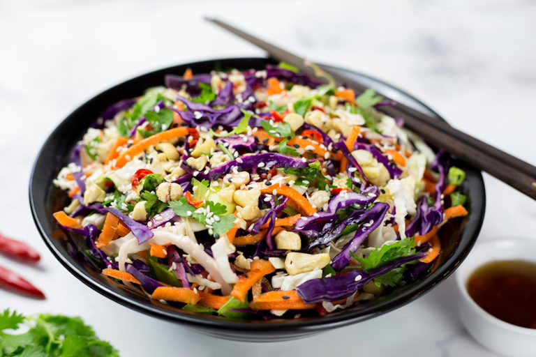 Asian slaw in black bowl with asian style dressing, chillis and coriander beside it on a white background.