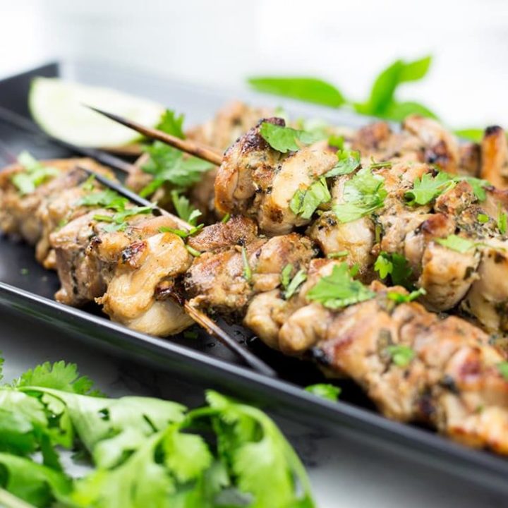 Thai chicken skewers with lemon wedge behind and coriander in front on black platter.
