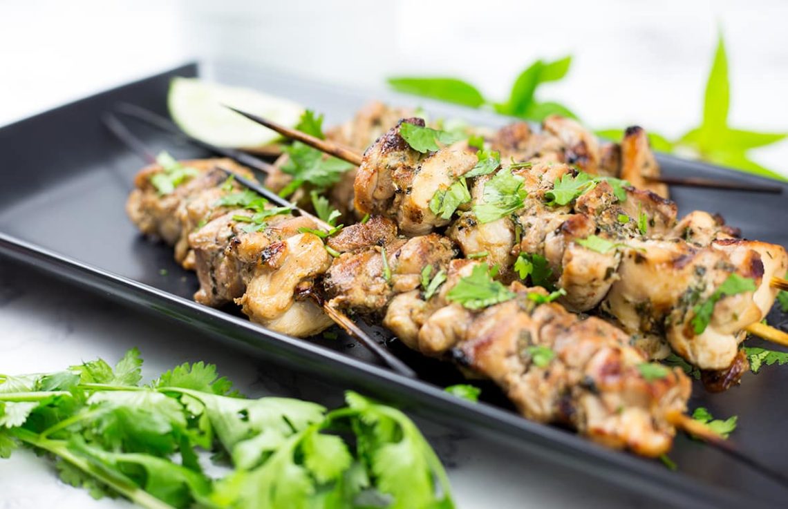 Thai chicken skewers with lemon wedge behind and coriander in front on black platter.