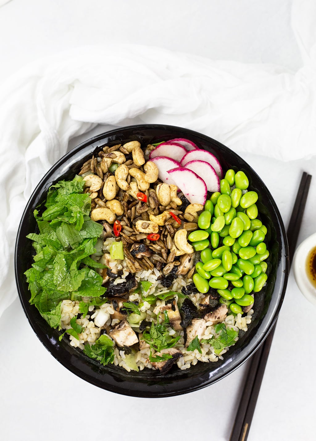 Birdseye view of Asian rice bowl with chopsticks, dressing to the right and white cloth behind.