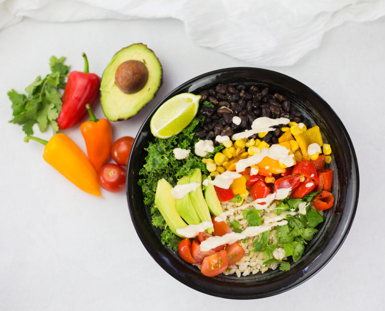 Birdseye view of vegan burrito bowl with avocado, red and orange pepper and coriander in background on a white backdrop.