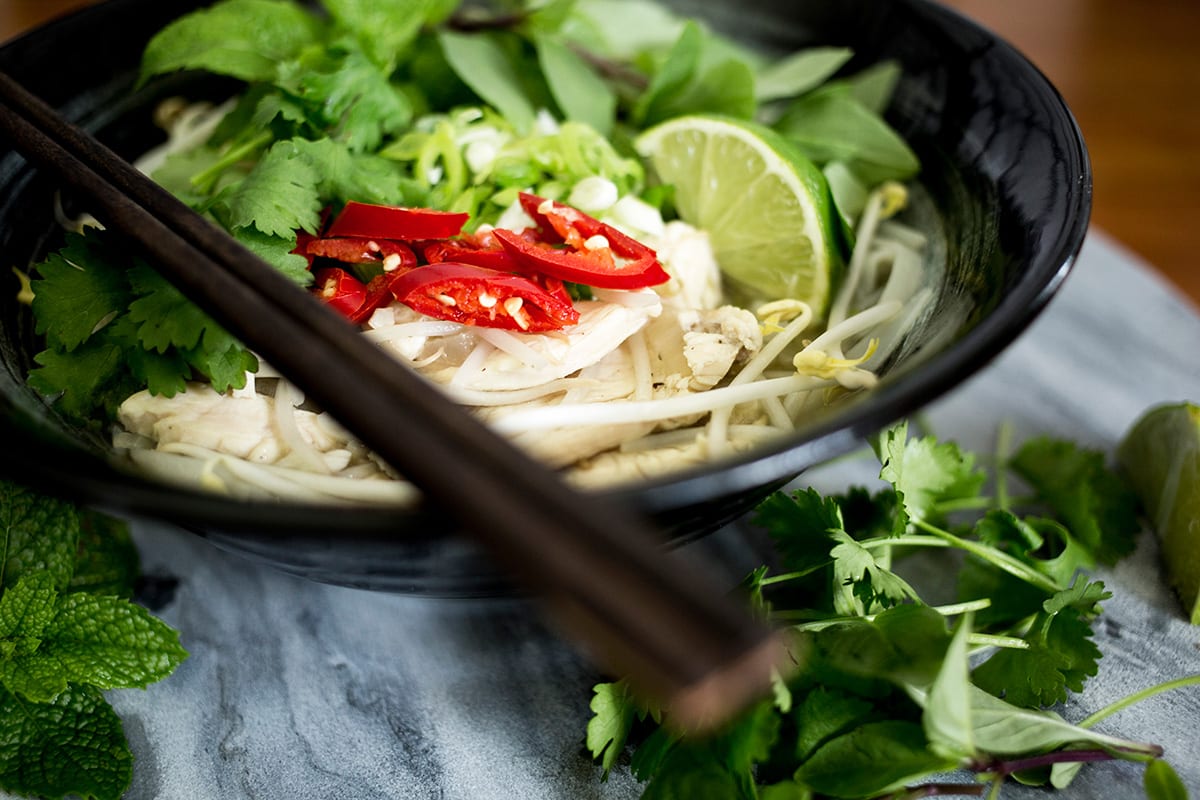 Close up of chicken pho in black bowl with chopsticks on the left on marble background.