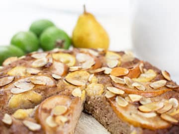 Close up of feijoa and pear cake with a slice removed on chopping board with fruit in background.