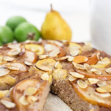 Close up of feijoa and pear cake with a slice removed on chopping board with fruit in background.