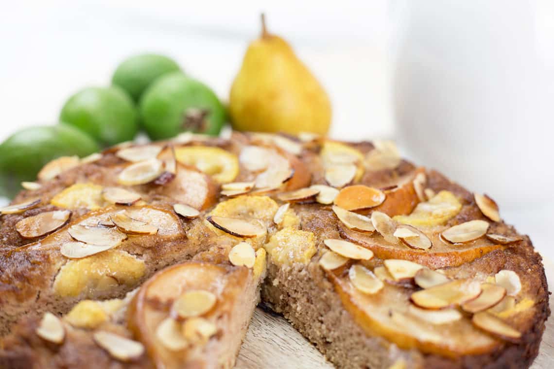 Close up of feijoa and pear cake with a slice removed on chopping board with fruit in background.