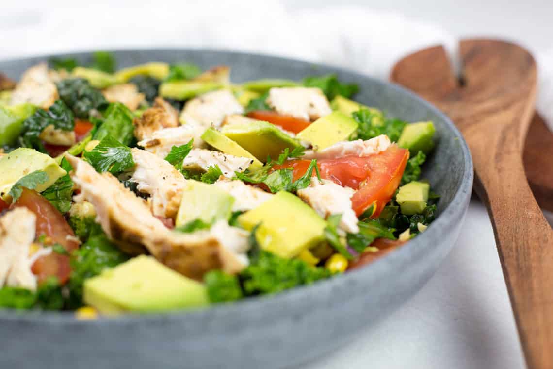 Close up of chicken salad on left with serving spoons on right, close up of chicken, avocado and tomatoes chopped in a bowl.