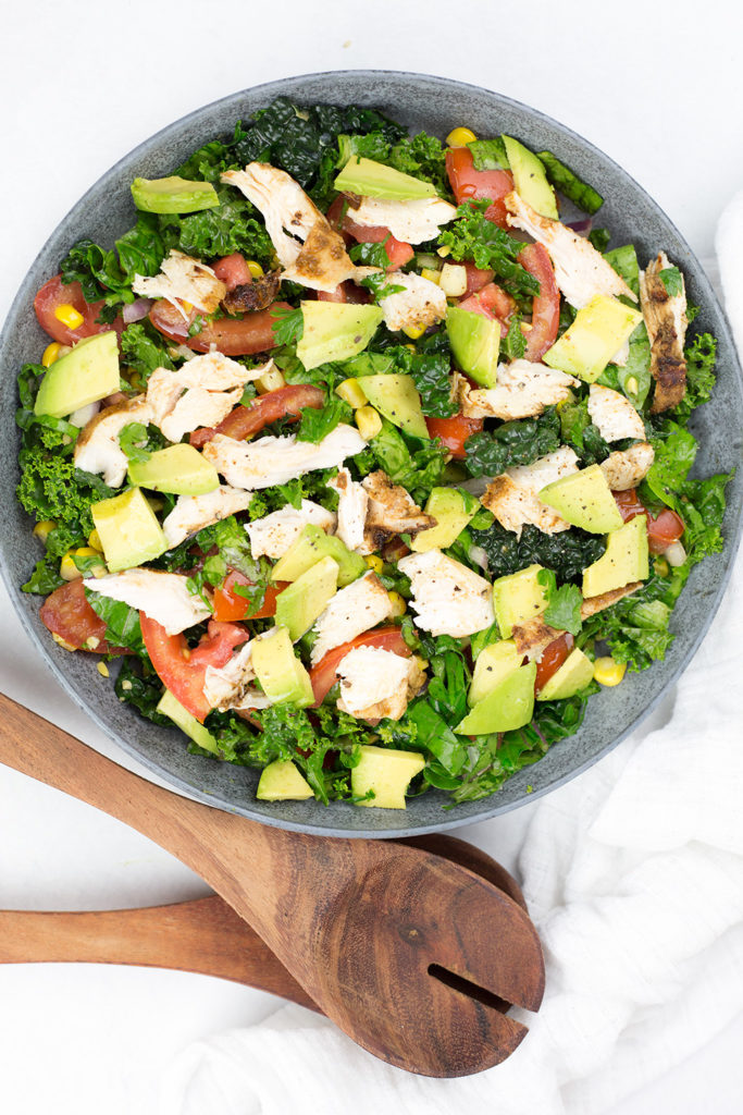 Close up of marinated chicken salad in blue/grey bowl with serving sppons. Shows lots of fresh vegetables such kale, avocado, tomato, corn.