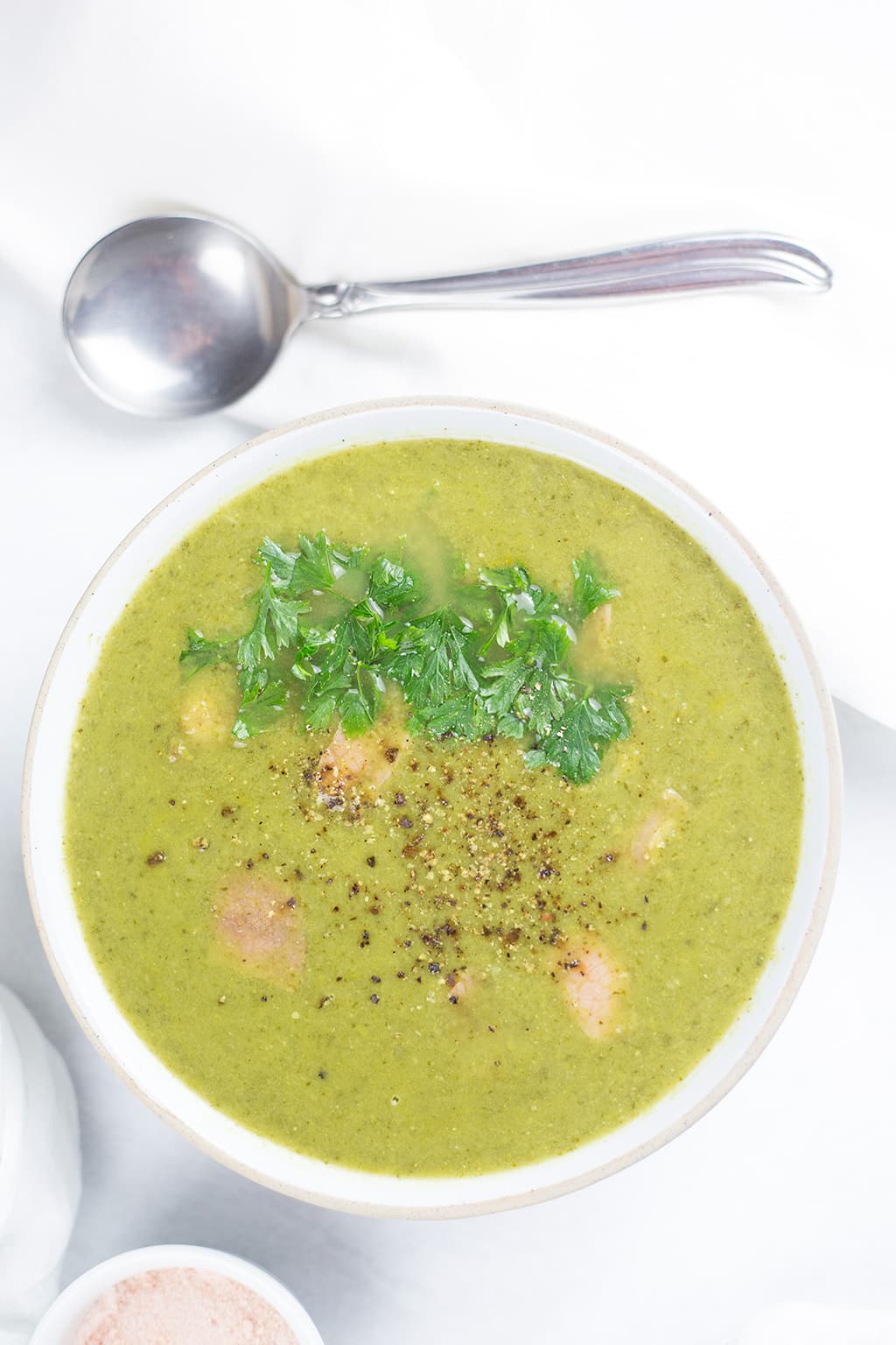 Birds eye view of pea and ham soup with parsley on top, silver spoon to the side and bowl of sea salt on other side.