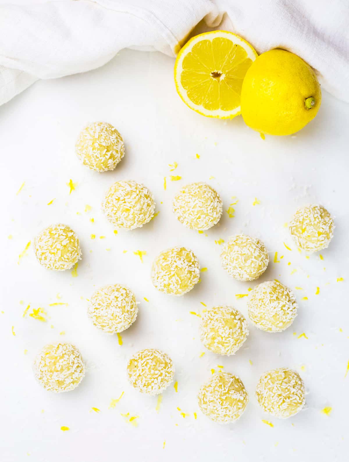 Birdseye view of lemon bliss balls scatted over white background with lemon zest in between and sliced lemon in background.