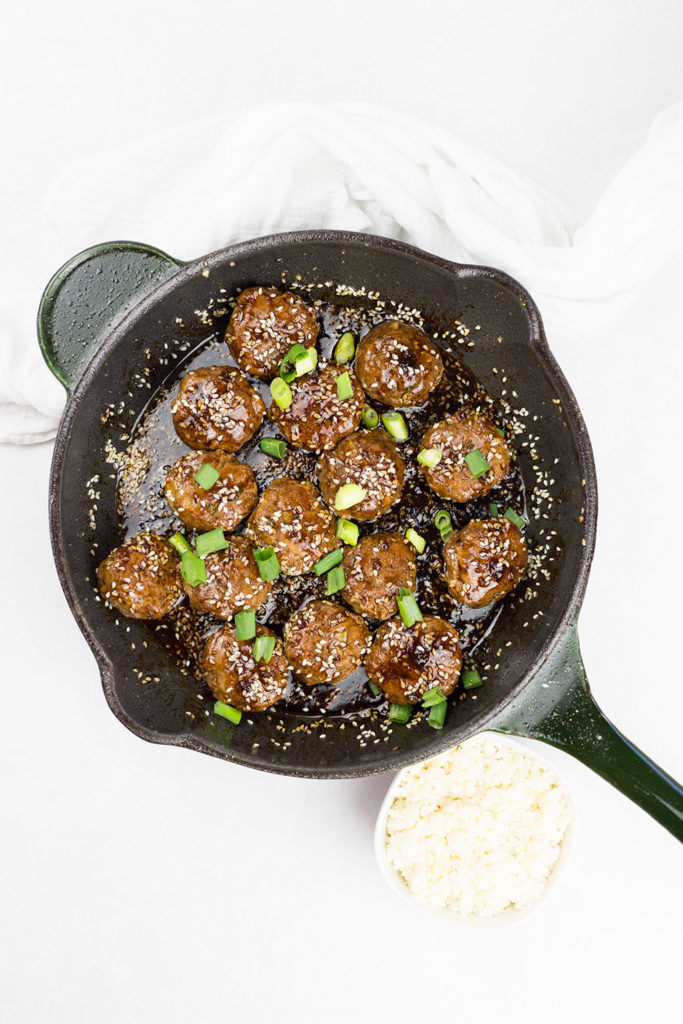 Birdseye view of Asian style meatballs in black skillet on white background with bowl of rice to the side.