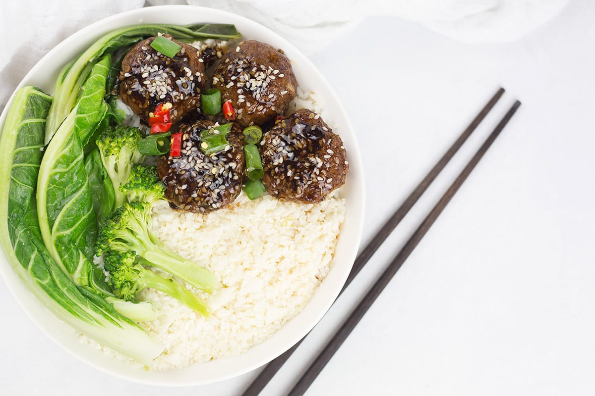 Stickey Asian meatballs in white bowl with rice and green vegetables and chopsticks to the side.