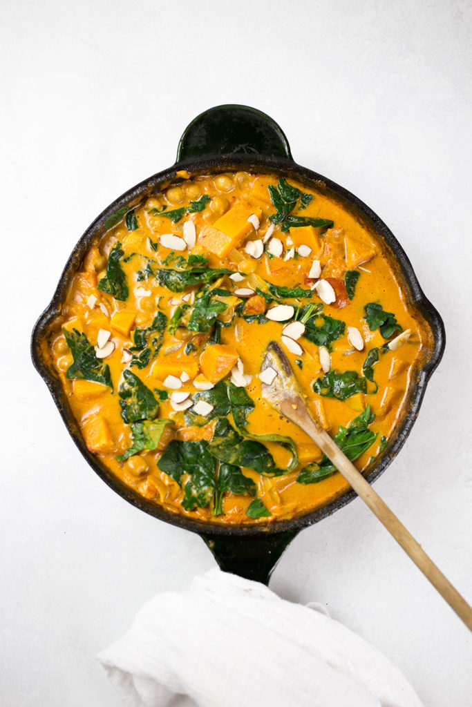 Birdseye view on butternut squash curry in black skillet with white cloth around handle. Process shot after the spinach and almonds have been added.