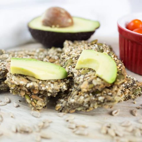 Close up of seed crackers with chopped avocado and red bowl of tomatoes in background.