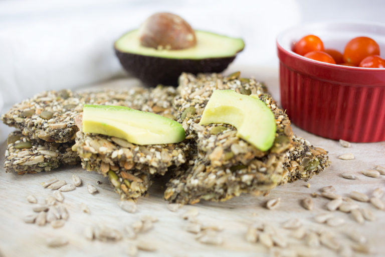 Close up of seed crackers with chopped avocado and red bowl of tomatoes in background.