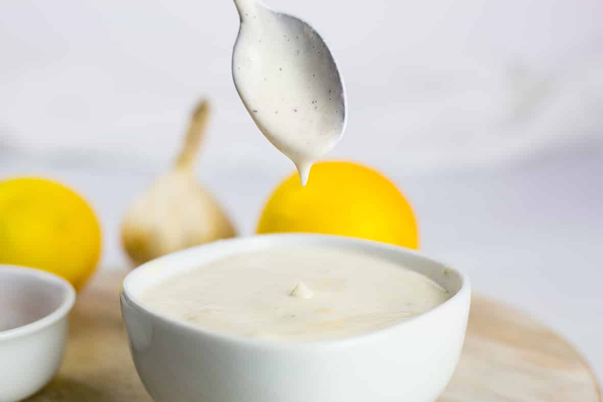 Lemon Tahini Dressing in a white bowl being dripped into bowl with a spoon, lemon and garlic in background.