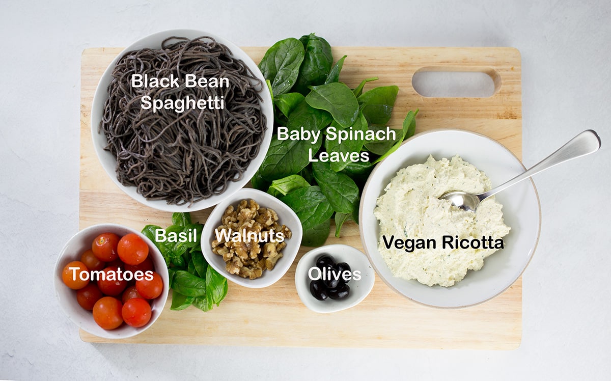 Spaghetti, tomatoes, spinach, olives, walnuts, basil, vegan ricotta on a chopping board.