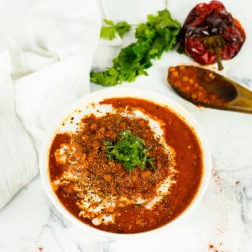 Birdseye view of red pepper and lentil soup with white cloth in the background and wooden spoon and a roasted red pepper and parsley.
