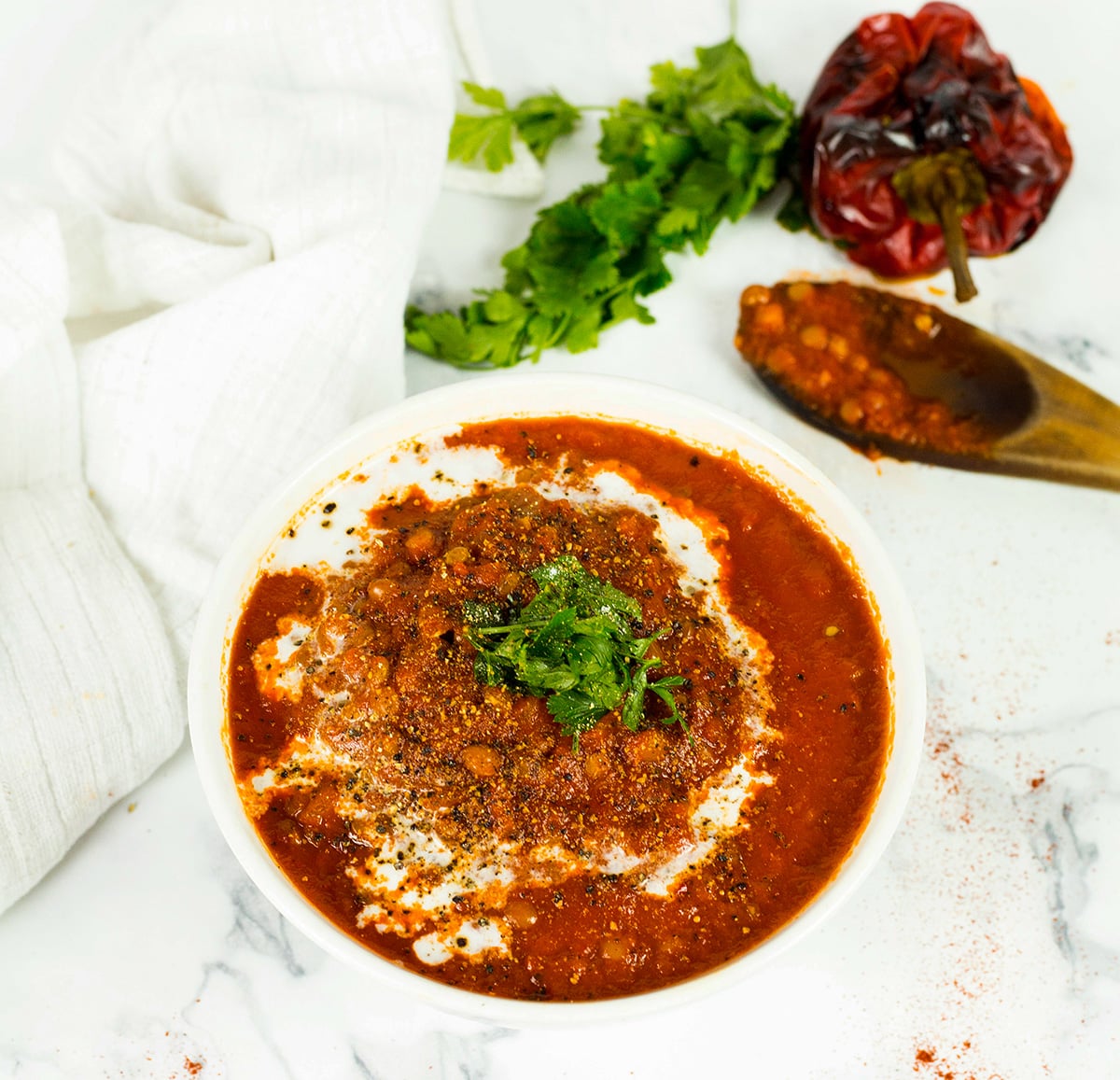 Birdseye view of red pepper and lentil soup with white cloth in the background and wooden spoon and a roasted red pepper and parsley.