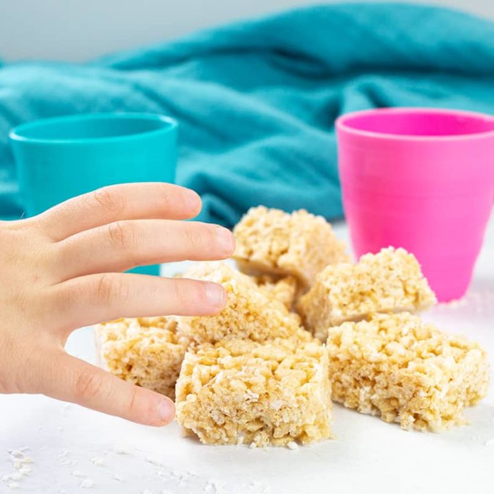 Childs hand reaching for rice bubble slice with pink and turquoise cup in background.