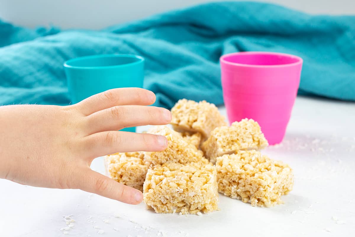 Childs hand reaching for rice bubble slice with pink and turquoise cup in background.
