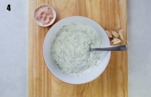 Tzaziki in white bowl on wooden background with garlic and salt to the side.