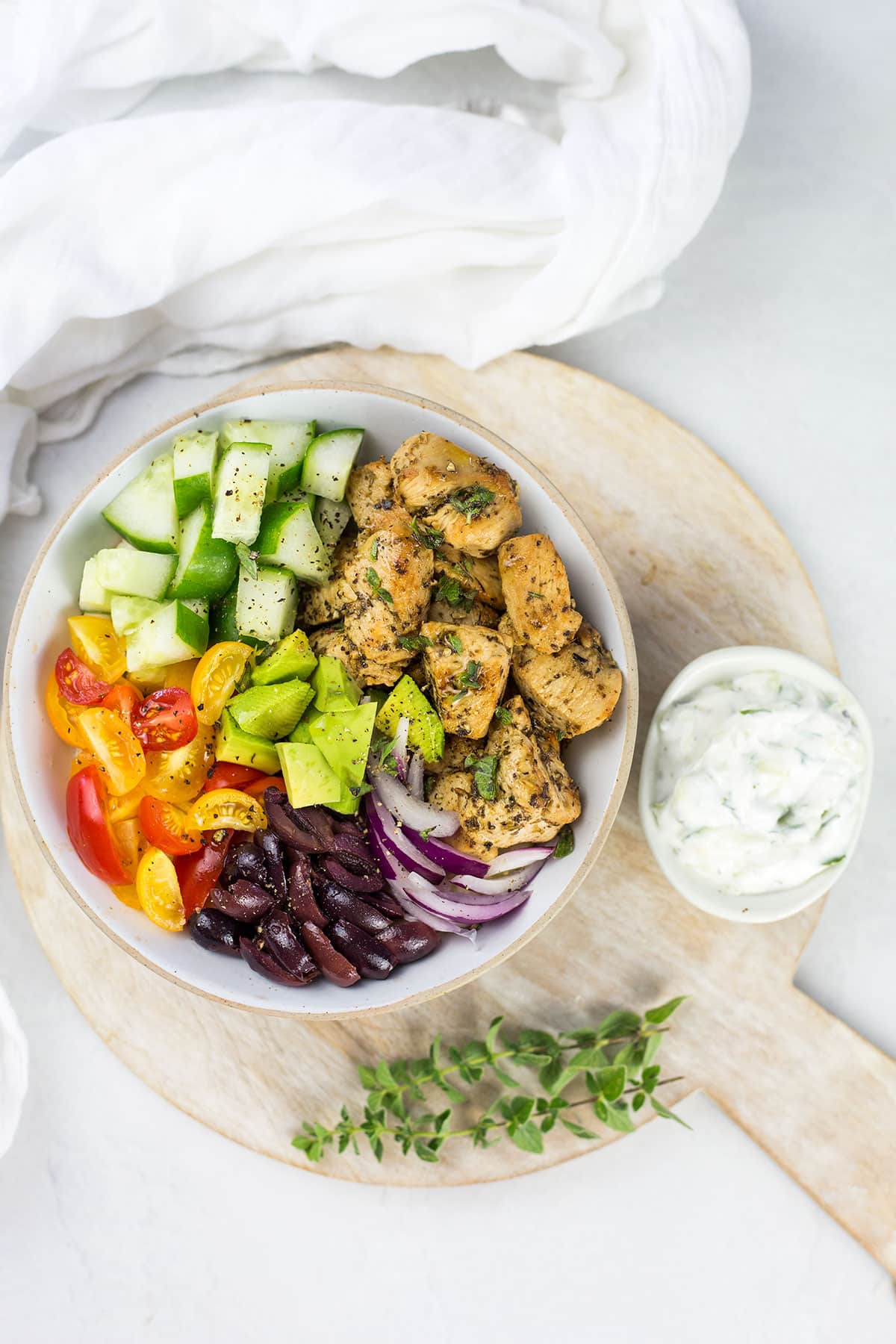 Birdseye view of Greek chicken bowl with tzatziki on wooden board and white cloth in background.