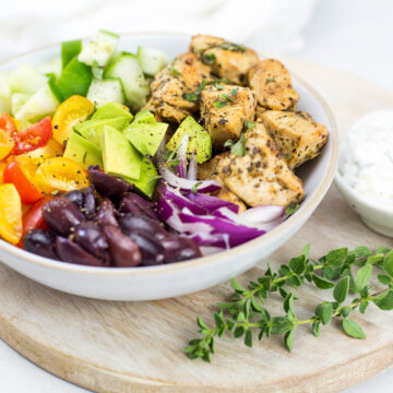 Greek chicken bowl on wooden board with fresh oregano to the side.