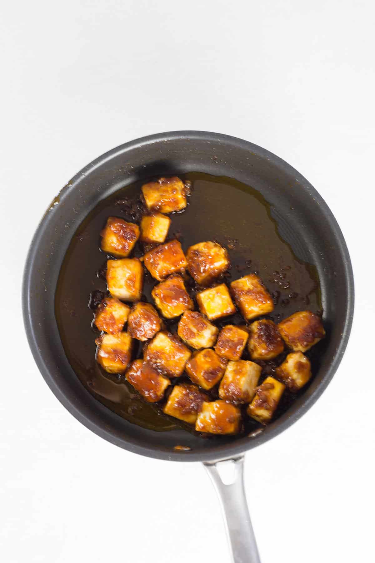 Coated tofu being cooked in sauce in black pan.