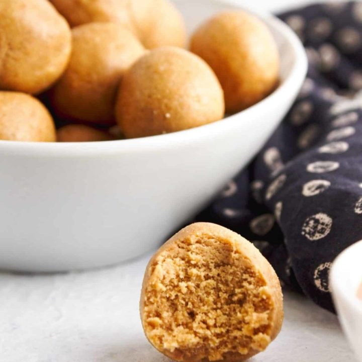 Bowl of peanut butteer protein balls with one half bitten on its own in front of bowl on white background.