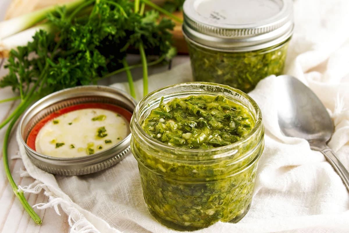 Coriander chimichurri in jars on white cloth with herbs and spoon in background.
