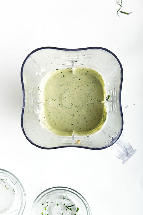 Blended ingredients for dairy free ranch dressing in belnder on white background. Empty glass bowls for herbs in front.