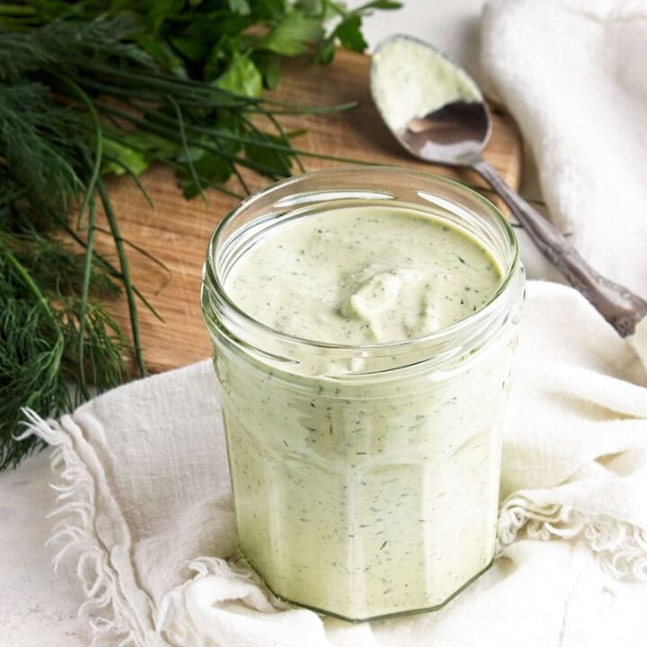 Dairy-free ranch dressing in glass jar with white cloth, teaspoon, fresh herbs and board behind.
