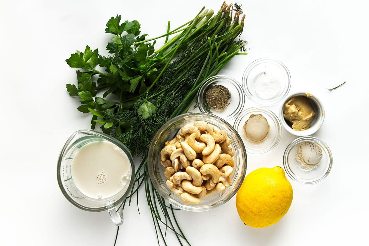 Ingredients for dairy free ranch dressing in glass bowls, jug on white background with sprig of fresh herbs and lemon beside.