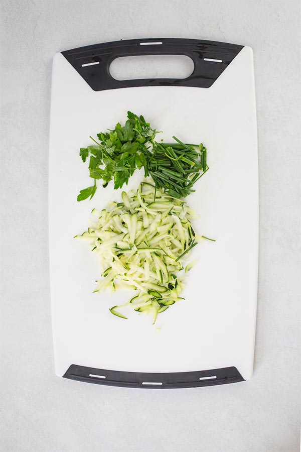 Grated zucchini and fresh herbs on chopping board.