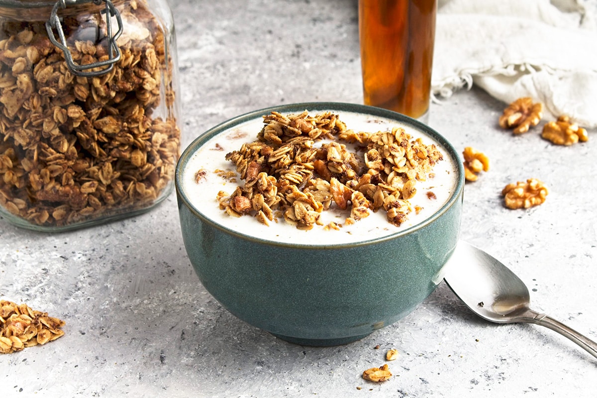 Maple Walnut Granola in blue bowl with spoon, jar and maple syrup behind.