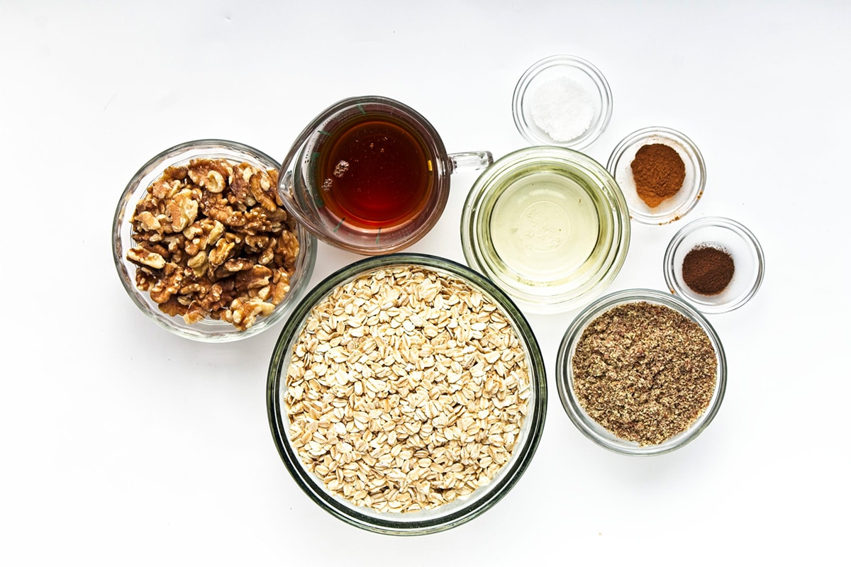 Ingredients for maple walnut granola in glass bowls and jug on white background.